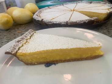 Ein Stück Zitronentarte mit goldgelber Füllung, bestäubt mit Puderzucker, liegt auf einem Teller. Dahinter sieht man ganze Zitronen und die Tarte.