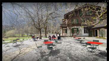 Eine Café-Szene im Freien mit roten Tischen und grünen Stühlen, umgeben von kahlen Bäumen. Die Leute sitzen da und genießen die Aussicht in der Nähe eines rustikalen braunen Hauses vor einer Bergkulisse.