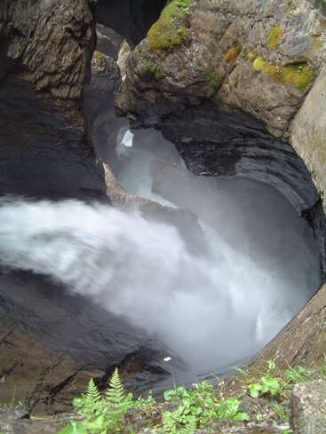 Ein mächtiger Wasserfall stürzt durch eine enge Felsschlucht. Moos, grüne Pflanzen und Farne klammern sich an die Felswände, während Nebel aus dem tosenden Wasser unten aufsteigt.