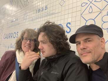 Three people pose together in front of a tiled wall with blue designs and text. The person in the middle eats a churro, everyone looks happy and relaxed.