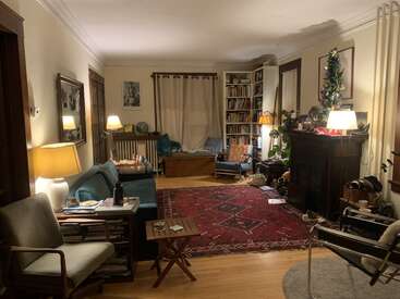 This cozy living room features warm lighting, a patterned red rug, teal sofa, bookshelves, armchairs, a decorated fireplace, plants, and a small table with snacks.