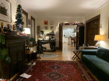 A cozy living room with a dark green sofa, red patterned rug, decorated Christmas tree, fireplace, piano, art, books, lamps, and kitchen in background.