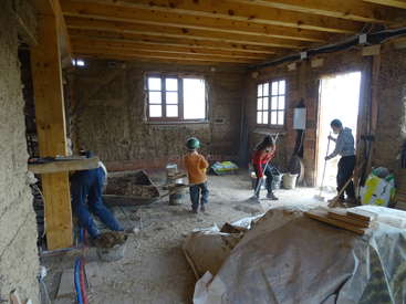 L'image représente un groupe d'enfants travaillant dans une pièce rustique avec des murs en briques apparentes, des poutres en bois et un sol en terre battue, entourés de matériaux de construction.