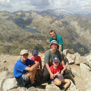 A imagem mostra uma família de cinco pessoas posando no cume de uma montanha rochosa, com uma vista deslumbrante de montanhas e lagos escarpados ao fundo.