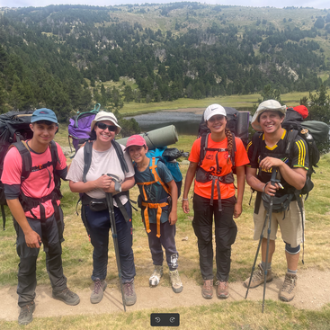 Cinco caminhantes, sorrindo e posando juntos, carregam mochilas e bastões de trekking. Eles estão ao ar livre em um caminho gramado com montanhas, floresta e um lago atrás deles.