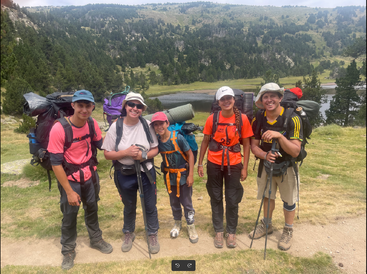 Five hikers, smiling and posing together, carry backpacks and trekking poles. They stand outdoors on a grassy path with mountains, forest, and a lake behind them.