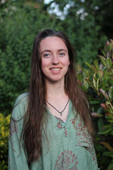 A young woman with long brown hair smiles at the camera. She wears a light green patterned shirt and a necklace, standing in a lush green garden.
