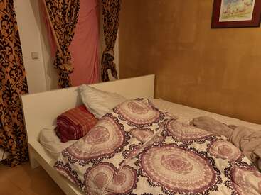 This image shows an unmade bed with patterned bedding, pillows, and a blanket. Ornate curtains and golden wall add warmth, with a framed painting above. Cozy atmosphere.