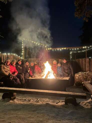 A group of people bundled in winter clothing sit around a warm, glowing campfire at night, surrounded by snow and twinkling string lights, enjoying togetherness.