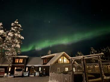 A cozy, snow-covered house glows warmly under a starry night sky, while vibrant green Northern Lights beautifully illuminate the wintry landscape and surrounding trees.