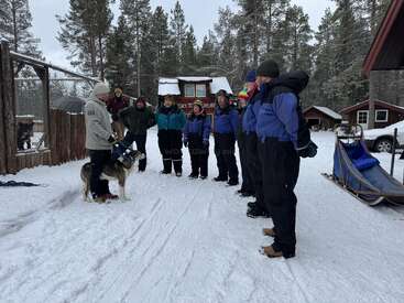 A group of people dressed in winter clothing stands in the snow, listening to a guide with a dog near a sled, surrounded by cabins and trees.