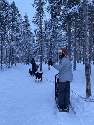 A person stands on a dog sled, smiling and waving amid a snowy forest. Several huskies pull the sled while trees covered in snow surround the scene.