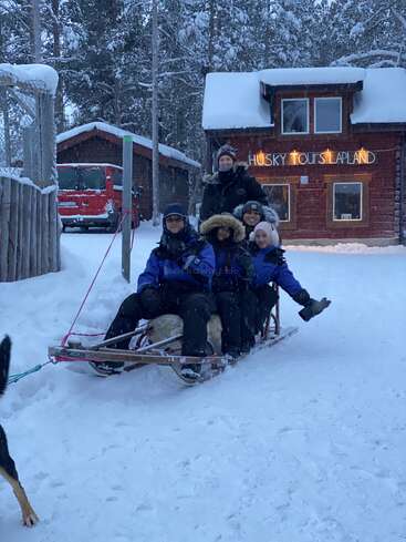 Four people bundled in winter gear sit on a wooden dog sled in snowy Lapland, outside a cozy cabin labeled "Husky Tours Lapland,” surrounded by fir trees.
