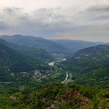 Un frondoso valle verde se extiende entre imponentes montañas, con un río serpenteante que atraviesa pequeños pueblos bajo un cielo nublado. Las onduladas colinas crean un paisaje tranquilo.
