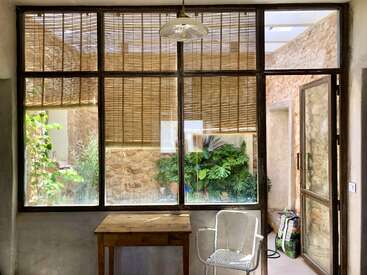 Una habitación iluminada por el sol, con paredes de piedra rústica y grandes ventanales cubiertos por persianas de bambú. Dentro, hay una mesa de madera, una silla blanca de metal y exuberantes plantas verdes en el exterior.