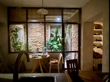 In einem gemütlichen Innenraum gibt es einen kleinen Tisch, Stühle und eine Spüle. Große Fenster geben den Blick frei auf einen Innenhof mit Steinmauern und üppigen grünen Pflanzen.