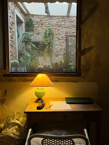 Ein gemütlicher Schreibtisch mit einer grünen Lampe, einem Notizbuch und einem Stift steht am Fenster. Draußen wachsen Kakteen und Weinreben an rustikalen Steinmauern. Ein einladender Arbeitsbereich.