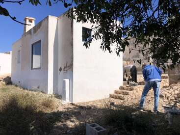 Zwei Personen arbeiten vor einem kleinen, weißen, rechteckigen Gebäude mit sichtbarer Abnutzung. In der Umgebung gibt es trockenes Gras, Steinstufen und schattenspendende Äste.