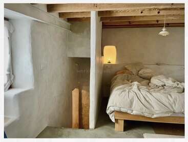 Ein gemütliches, minimalistisch eingerichtetes Schlafzimmer mit sichtbaren Holzbalken, rustikal verputzten Wänden, einem ungemachten Bett mit neutraler Bettwäsche, warmer Beleuchtung und einer kleinen Treppe, die neben dem Bett teilweise zu sehen ist.