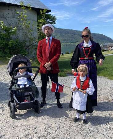 A family stands outdoors in traditional Norwegian attire. A man in a red jacket, woman in black dress, two children—one holding a Norwegian flag, one in a stroller.