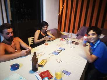 A imagem mostra uma família de três pessoas sentadas à mesa, jogando cartas, com bebidas e lanches espalhados, em um ambiente doméstico aconchegante.