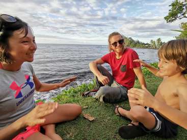 A imagem mostra uma família de três pessoas sentadas na grama perto do oceano, com a mulher e o homem conversando enquanto o menino gesticula com entusiasmo.