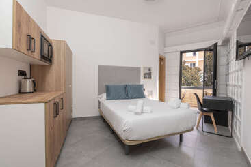 Ein modernes Studio-Apartment mit einem Doppelbett, einer Küchenzeile, einem Schreibtisch und einem Stuhl. Ein großes Fenster bietet natürliches Licht und einen Blick auf die Bäume und Gebäude draußen.