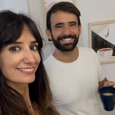 Eine Frau und ein Mann lächeln in einem Haus. Der Mann hält einen blauen Becher in der Hand. An der weißen Wand hinter ihnen sind zwei gerahmte Kunstwerke zu sehen.