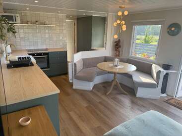 This image shows a cozy, modern kitchen and dining nook with a round table, built-in seating, stylish lighting, green cabinets, wooden countertops, and large window views.