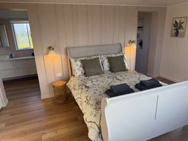 A cozy bedroom with a white wooden bed, floral bedding, green accent pillows, bedside lamps, wooden floors, soft lighting, and an adjacent bathroom visible. Stylish, inviting, serene.
