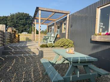 A cozy outdoor patio features a light blue picnic table, gravel ground, wooden decking with planters, shaded seating area, and gray paneled walls under clear skies.