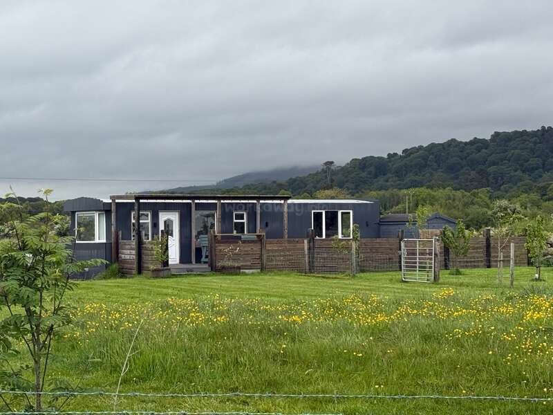 A modern, single-story house sits amidst a grassy field with yellow flowers. Wooden fencing surrounds the property. Green hills and cloudy skies are in the background.