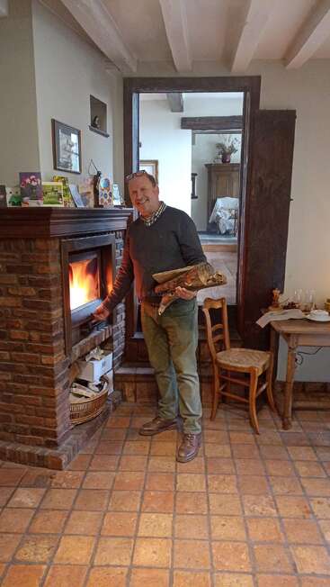 A man stands by a cozy brick fireplace, holding logs and smiling. The homey room features rustic furniture, cards on the mantel, and warm terracotta tiles.