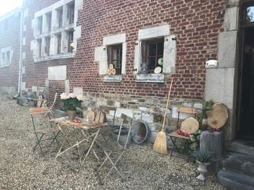 Une scène rustique en plein air avec une table et des chaises patinées par le temps, des fleurs, des bûches, des pommes, un balai et une décoration vintage, le tout devant une maison en briques aux petites fenêtres.