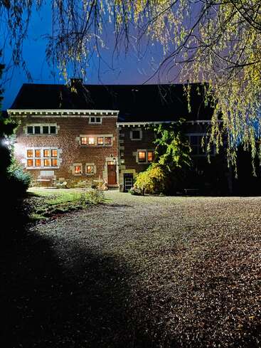 Ein Backsteinhaus mit leuchtenden Fenstern steht in der Abenddämmerung umgeben von Bäumen. Durch helle Lichter beleuchtet, wirken sein Garten und der Backsteinweg einladend und ruhig.
