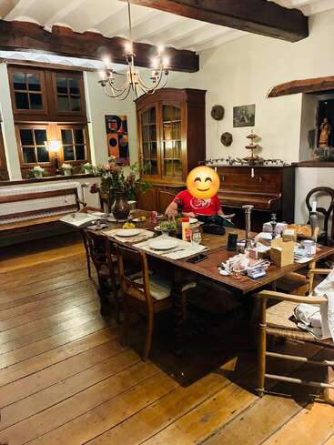 Ein gemütliches Esszimmer mit Holzböden und Holzbalken, einem großen gedeckten Tisch, Blumen und einer lächelnden Person mit einem Emoji-Gesicht am Tisch.