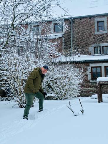 Eine Person in einer grünen Jacke und einem Hut steht im tiefen Schnee vor einem Backsteinhaus. Schneebedeckte Bäume umgeben die gemütliche Winterszene und schaffen eine friedliche Atmosphäre.