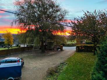 Ein krasser Sonnenuntergang macht den Himmel bunt und taucht alles in warme Orange- und Rosatöne. Bäume mit Herbstlaub umrahmen eine Auffahrt, ein Auto, einen Anhänger und eine ruhige Landschaft.
