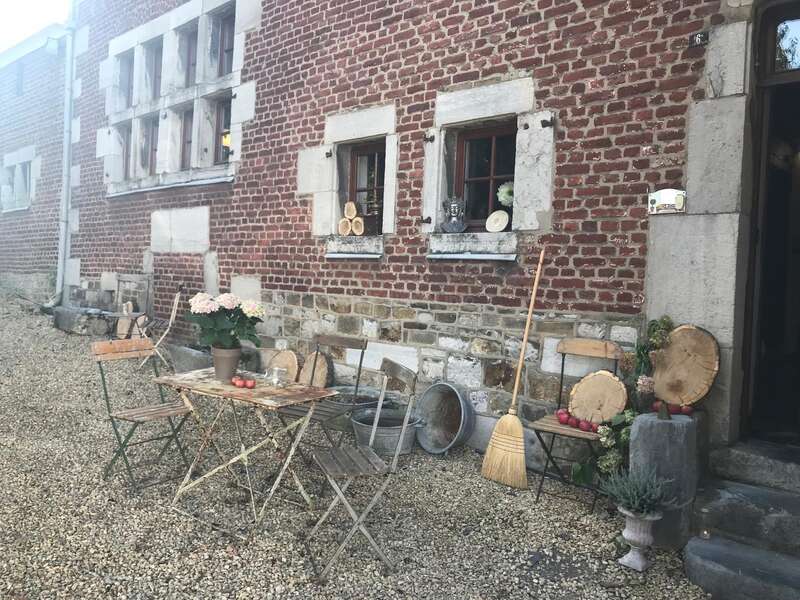 A rustic outdoor scene with a weathered table and chairs, flowers, logs, apples, a broom, and vintage decor set against a brick house with small windows.