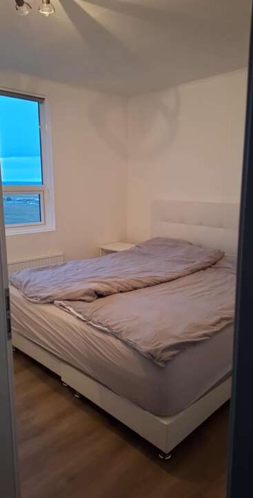 Ein kleines, minimalistisches Schlafzimmer mit einem großen Bett, hellgrauer Bettwäsche, weißem Kopfteil, Holzboden, einem einzelnen Fenster und sanfter Beleuchtung schafft eine gemütliche, friedliche Atmosphäre.