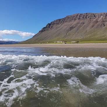 As ondas batem suavemente em uma praia clara, com campos verdes distantes e edifícios espalhados sob uma montanha imponente e escarpada, sob um céu azul brilhante. Cena tranquila.