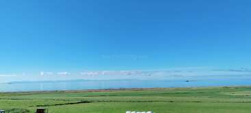 Un paysage serein avec un ciel bleu clair, des montagnes lointaines, une eau calme et de vastes champs verdoyants. La scène semble paisible et étendue, suggérant un environnement calme et naturel.