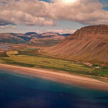 Un sereno paisaje costero con campos de un verde vibrante, una costa arenosa, aguas de un azul intenso, montañas escarpadas, nubes dispersas y parcelas de tierra de cultivo bajo un cielo brillante.