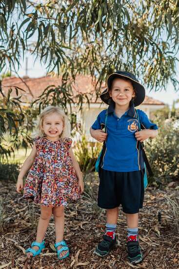 Two smiling children stand outdoors under a tree. The girl wears a colorful dress, and the boy, in a school uniform and hat, carries a backpack.