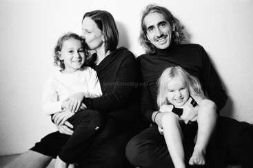 A black-and-white photo of a smiling family of four sitting against a wall. The parents hold their two young children, radiating warmth, happiness, and love.