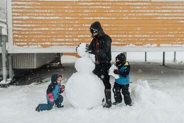 Un adulte emmitouflé et deux enfants construisent ensemble un bonhomme de neige par temps de neige, avec un bâtiment en bois jaune à l'arrière-plan et de la neige recouvrant le sol.