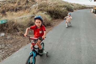 Deux jeunes enfants font du vélo sur un chemin pavé entouré d'herbe sèche. L'enfant à l'avant porte une chemise rouge et un casque, et sourit avec assurance à l'appareil photo.