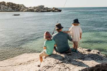 Un homme et deux enfants sont assis sur une côte rocheuse et s'amusent à pêcher ensemble. Le soleil brille, la mer est calme et ils portent des chapeaux et des vêtements d'été.