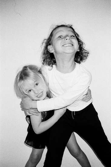 Deux enfants souriants, une fille en robe et un garçon en chemise à manches longues, se serrent chaleureusement dans les bras sur un fond blanc uni, affichant des expressions joyeuses.