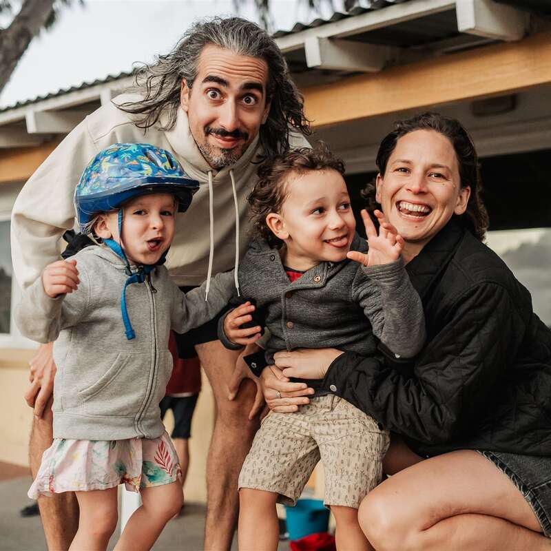 A joyful family of four poses outside, smiling and making playful expressions. The children wear casual clothes, one in a helmet, while the parents happily embrace them.